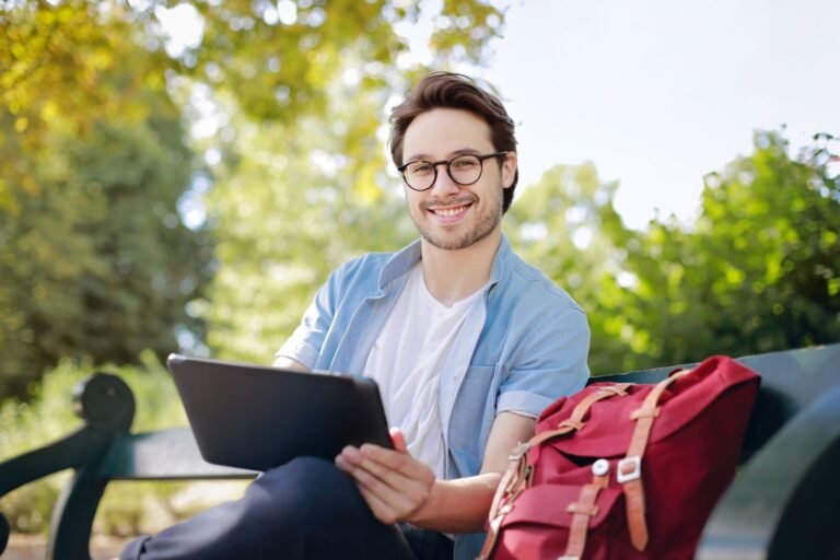 Smiling young man with glasses using a tablet on a park bench with a red backpack nearby.